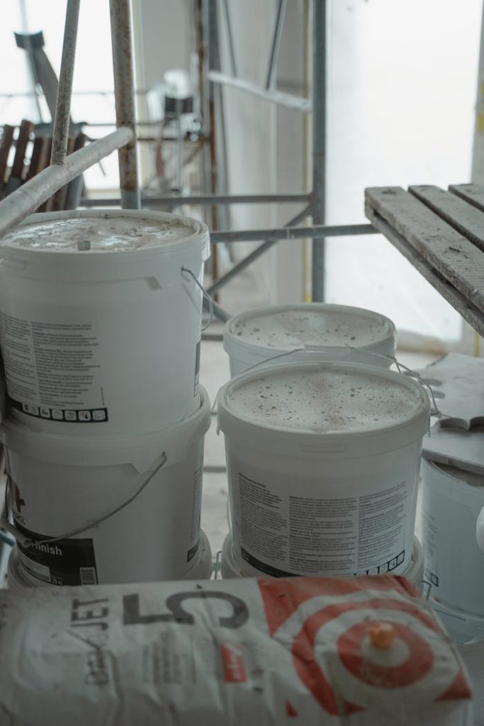 Several white paint buckets stacked in an indoor construction area with scaffolding.